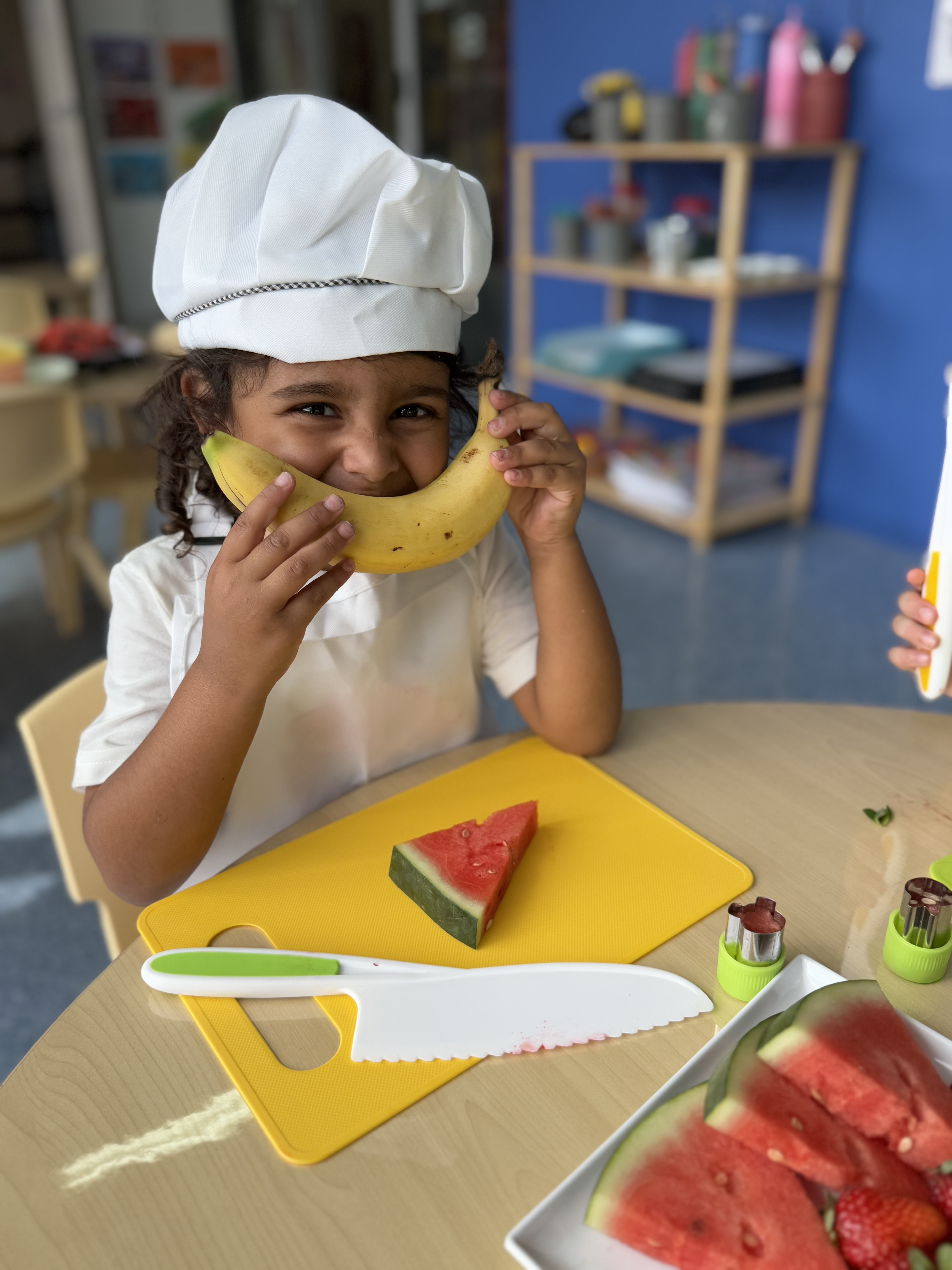 Young girl in a chef hat playfully holding a banana in front of her mouth, smiling.